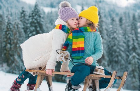 Foto de Los niños se besan. Niño y niña disfrutando de un paseo en trineo. Los niños se besan juntos, juegan al aire libre en la nieve en las montañas en invierno. Niños Vacaciones de Navidad - Imagen libre de derechos