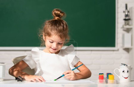 Téléchargez les photos : Enfant fille dessine dans la salle de classe assis à une table, s'amusant sur fond de tableau noir de l'école. enfant élève apprendre la peinture - en image libre de droit