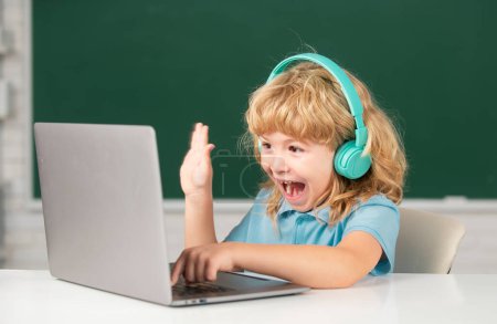 Téléchargez les photos : Écolier excité, élève étonné porte casque écrit sur ordinateur portable, écouter leçon audio utiliser ordinateur. Videocall, e-étude avec tuteur à distance - en image libre de droit