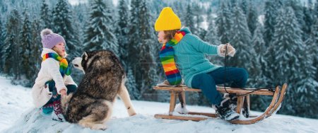 Téléchargez les photos : Enfant garçon et petite fille avec chien husky profitant d'une balade en traîneau. Les enfants font de la luge ensemble, jouent dehors dans la neige sur les montagnes en hiver. Enfants frère et sœur en vacances de Noël - en image libre de droit