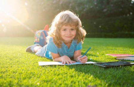 Foto de Hermoso niño escribiendo notas en copybook, acostado en la hierba en el fondo del prado. Niños leyendo libro en el parque - Imagen libre de derechos