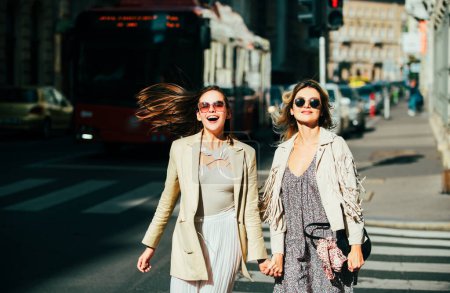Photo for Pretty girls walking on street outdoors and having fun. Best girlfriends, urban city style. Two friends laughing and walking in the city centre, wearing sunglasses - Royalty Free Image