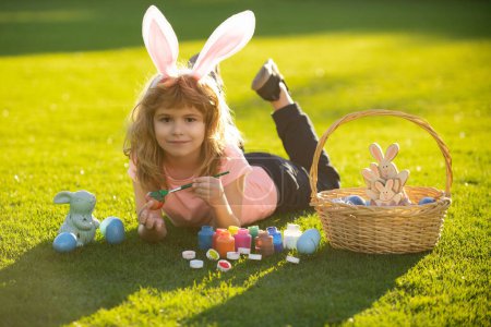 Foto de Kid laying on grass in park wit easter eggs. Children celebrating easter painting eggs. Kid in rabbit costume with bunny ears outdoor - Imagen libre de derechos