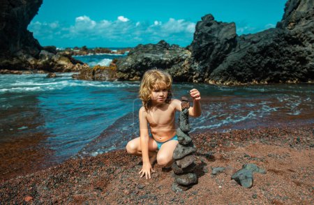 Foto de Niño lindo niño haciendo pirámide de piedras de mar en la playa. Balance de vida y concepto de armonía. Equilibrar la naturaleza. Niño jugando con piedras en la playa - Imagen libre de derechos