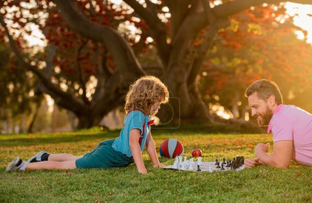Téléchargez les photos : Père et fils jouant aux échecs allongés sur l'herbe à Lawn Park. Fête des Pères, famille amoureuse, parentalité, concept d'enfance. Enfant apprenant à jouer aux échecs. Petit garçon pense ou planifie le jeu d'échecs. Échec mat - en image libre de droit
