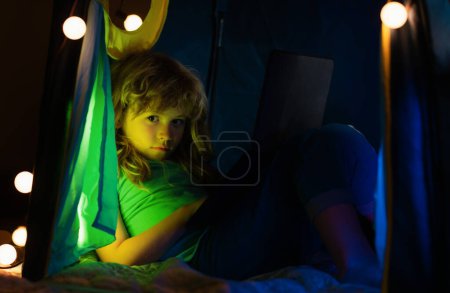 Foto de Niño pequeño usando el ordenador portátil, sentado en la tienda acogedora oscura en casa. Niños listos aprendiendo en casa. Niño niño viendo dibujos animados en el dormitorio por la noche - Imagen libre de derechos