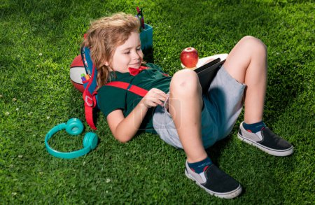 Foto de Niño sentado en la hierba con la tableta en el parque. Escuela al aire libre. Sonriente chico mantenga la tableta de ordenador. Escuela, educación, aprendizaje, tecnología ocio concepto al aire libre - Imagen libre de derechos