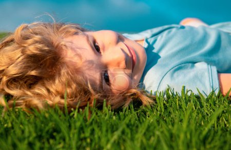 Téléchargez les photos : Portrait d'un petit garçon souriant allongé sur de l'herbe verte et rêvant. Enfant mignon profitant de la nature en plein air. Enfant en bonne santé jouant dehors dans un parc d'été sur une pelouse - en image libre de droit