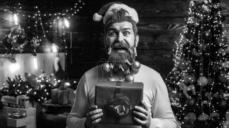 Téléchargez les photos : Des émotions cadeaux. Père Noël excité avec un cadeau. Portrait d'homme barbu avec barbe décorée et chapeau de Père Noël. Styling Santa hipster avec une longue barbe posant sur le fond en bois de Noël - en image libre de droit