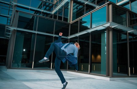 Téléchargez les photos : Homme d'affaires excité gardant les bras levés et exprimant sa positivité tout en restant debout à l'extérieur avec un immeuble de bureaux - en image libre de droit