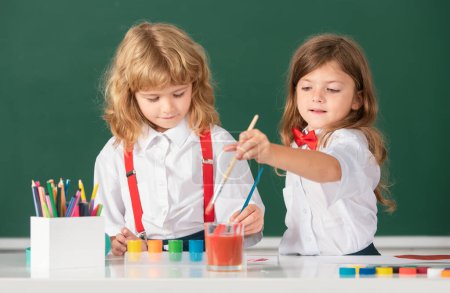 Téléchargez les photos : Éducation de la petite enfance. Peinture écolière fille et garçon avec peinture couleur et pinceau en classe - en image libre de droit