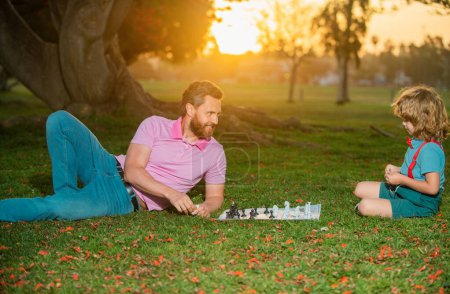 Téléchargez les photos : Joyeux famille en plein air. Père et fils jouent aux échecs dans le jardin d'été. Le petit garçon joue aux échecs avec ses parents. Développement cognitif. Jeu intellectuel pour enfants. Enfants intelligents - en image libre de droit