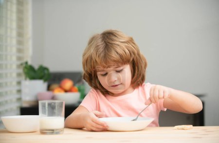 Foto de Niño pequeño caucásico comiendo sopa saludable en la cocina. Primeros planos de lindo niño comiendo sabrosas gachas o cereales con leche en casa, sentado en la mesa en la cocina - Imagen libre de derechos
