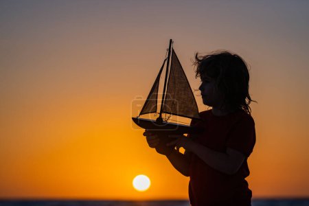 Téléchargez les photos : Silhouette d'enfant jouant avec jouet bateau à voile sur la mer coucher du soleil. Petit garçon jouant avec un voilier jouet, un bateau jouet. Concept voyage et aventure. Enfant se sentant aventureux pendant la croisière - en image libre de droit