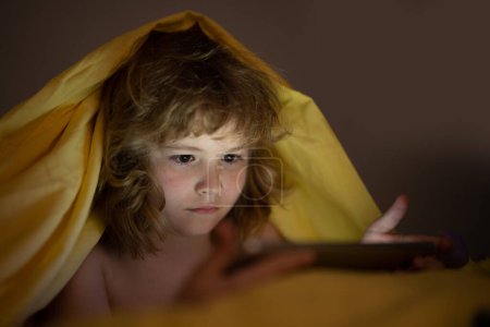 Foto de Niño en la cama debajo de una manta juega en una tableta en un juego en la oscuridad. La cara de los niños está iluminada por una tableta. Niño usando la tableta en la cama en casa. Niño en el dormitorio viendo películas o jugando - Imagen libre de derechos