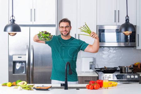 Téléchargez les photos : Homme dans la cuisine moderne, préparant des aliments sains seul, cuisine salade. Beau homme prépare une salade végétalienne fraîche dans la cuisine à la maison. Une alimentation saine est une vie saine - en image libre de droit