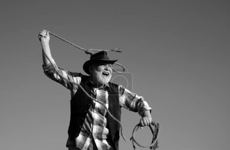 Photo for Senior western cowboy throwing lasso rope. Bearded wild west man with brown jacket and hat catching horse or cow. Rodeo or ranch - Royalty Free Image