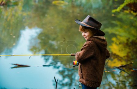 Téléchargez les photos : Enfant apprenant à pêcher, tenant une canne sur un lac. Enfant avec canne à pêche. Petit pêcheur dans un chapeau - en image libre de droit