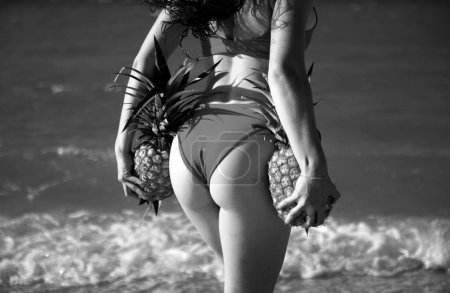 Téléchargez les photos : Femme fesses avec ananas en maillot de bain bikini sur sable mer - en image libre de droit