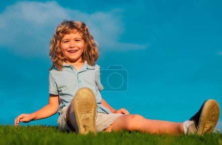 Téléchargez les photos : Garçon enfant heureux assis sur l'herbe verte à l'extérieur dans le parc d'été - en image libre de droit