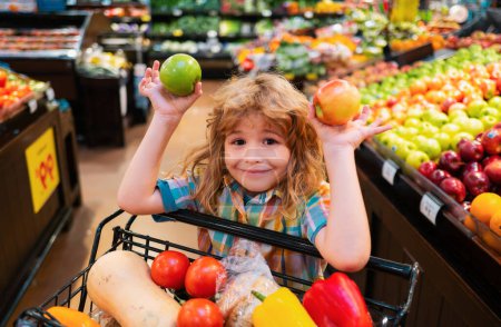 Téléchargez les photos : Enfant choisissant une pomme dans un magasin. Petit enfant drôle sourire et tenant pomme. Achats d'enfants au supermarché. Petit garçon choisissant pomme fraîche dans le magasin local - en image libre de droit