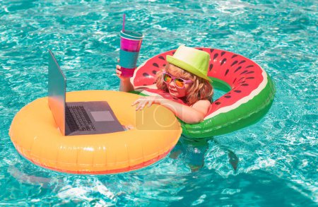 Téléchargez les photos : Travail en ligne et se détendre dans la piscine. Enfant se détendre dans la piscine, en utilisant un ordinateur portable dans l'eau d'été. Enfant étude en ligne ou de travailler sur la plage de la mer tropicale. Concept de technologie pour la vie - en image libre de droit