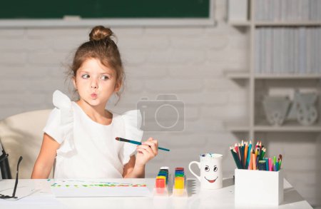Téléchargez les photos : Enfant fille dessine dans la salle de classe assis à une table, s'amusant sur fond de tableau noir de l'école. Apprentissage de l'enfance, compétences artistiques des enfants - en image libre de droit