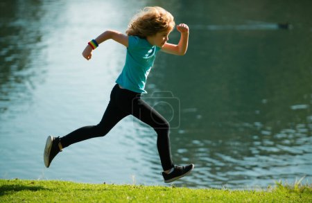 Téléchargez les photos : Enfant garçon courir ou faire du jogging près du lac sur l'herbe dans le parc. Garçon sportif coureur courir dans le parc d'été. Des enfants actifs, des enfants sportifs. Le jogging aide le corps à être fort - en image libre de droit