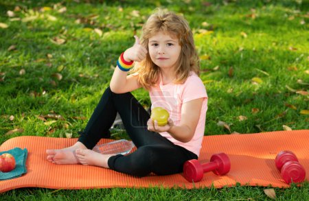 Foto de Estilo de vida saludable y concepto de comida saludable. Niño pequeño en ropa deportiva comiendo manzana sentado en la alfombra deportiva después de entrenar en el soleado día de primavera - Imagen libre de derechos