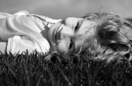 Téléchargez les photos : Portrait d'un petit garçon souriant allongé sur de l'herbe verte et rêvant. Enfant mignon profitant de la nature en plein air. Enfant en bonne santé jouant dehors dans un parc d'été sur une pelouse - en image libre de droit