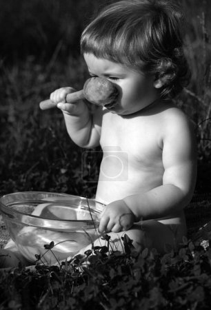 Téléchargez les photos : Bébé drôle assis sur l'herbe tenant cuillère en bois pour la nourriture - en image libre de droit