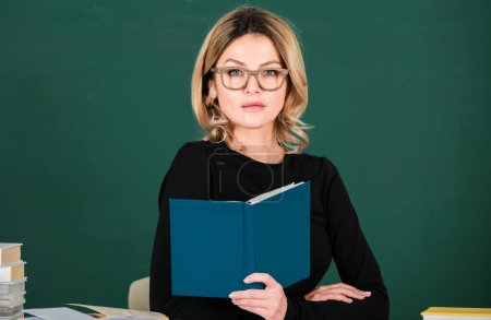 Foto de Modern attractive teacher. Portrait of smart young woman in glasses with book on the blackboard in class at high school or collage. Female student study in university. Study concept - Imagen libre de derechos