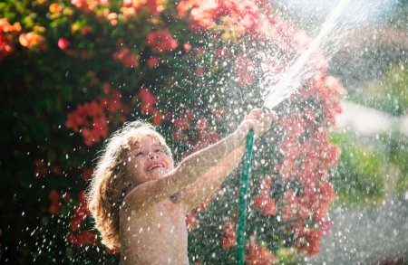 Téléchargez les photos : Petit enfant jardinier aidant à arroser les fleurs avec tuyau d'arrosage dans le jardin d'été. Travaux saisonniers - en image libre de droit