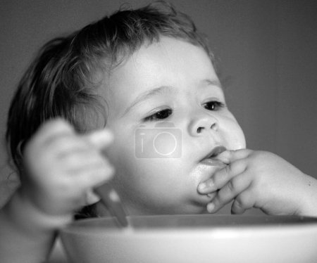 Foto de Lame sabrosos dedos. Retrato de niño caucásico lindo con cuchara. Bebé desordenado hambriento con plato después de comer puré - Imagen libre de derechos