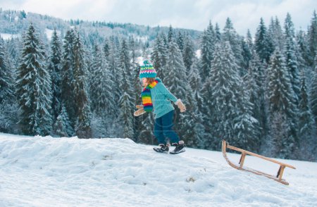 Téléchargez les photos : Boy kid sliding with sledge in the winter snow. Christmas kids holidays and Happy New Year. Snow covered trees in the mountains on winter landscape - en image libre de droit