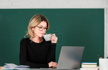 Photo for Portrait of teacher or female tutor working at table and drinking coffee in college or high school. Young women student studying in class - Royalty Free Image