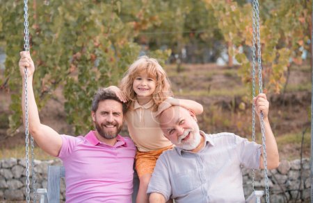 Foto de Tres generaciones de hombres juntos, retrato de un hijo sonriente, padre y abuelo balanceándose en el columpio, divirtiéndose en el parque al aire libre - Imagen libre de derechos