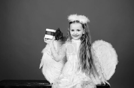 Téléchargez les photos : Enfant fille ange avec cadeau cadeau, portrait en studio. Petit ange aux ailes blanches tient cadeau, fée heureuse. Enfant ange mignon avec des ailes d'anges, isolé sur rouge. Saint Valentin - en image libre de droit
