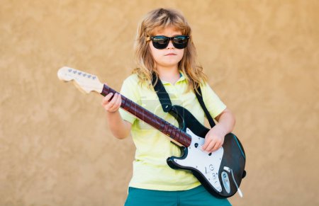 Téléchargez les photos : Child musician guitarist playing electric guitar. Funny child with blonde curly hair playing guitar on beige yellow background - en image libre de droit
