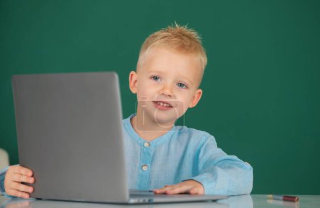 Téléchargez les photos : Enfant garçon utilisant un ordinateur portable à l'école. Cute pupille visage gros plan sur fond de tableau noir - en image libre de droit