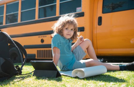 Foto de El alumno hace la tarea escolar tendido en el césped en el parque cerca del autobús escolar. Escuela niño al aire libre - Imagen libre de derechos