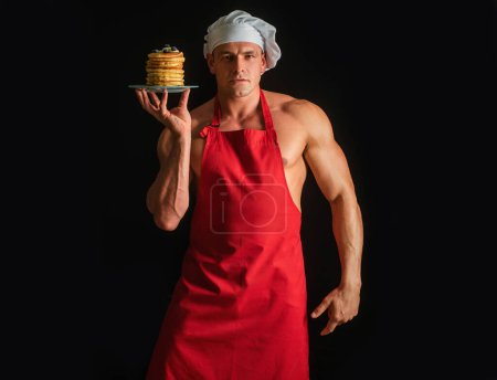 Foto de Hombre sexy en chefs sombrero con dulce pila casera de panqueques con jarabe aislado en negro. Chef hombre muscular con delantal hornear panqueques con bayas y jarabe de arce - Imagen libre de derechos