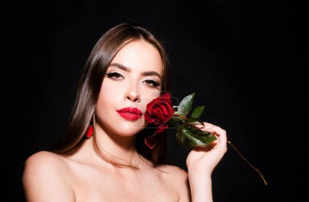 Foto de Brunette woman with red rose. Beautiful girl with reses flowers on a black background. Closeup face of young beautiful woman with a healthy clean skin and bright makeup - Imagen libre de derechos