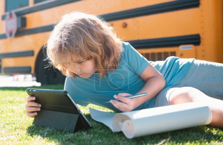 Foto de El niño hace la tarea escolar tendido en el césped en el parque cerca del autobús escolar. Escuela niño al aire libre - Imagen libre de derechos