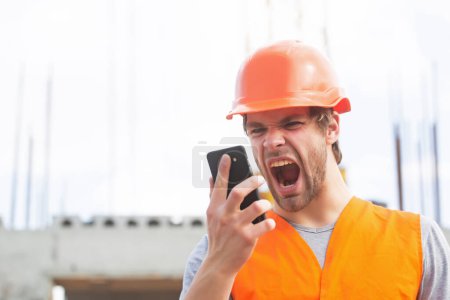 Téléchargez les photos : Homme constructeur travailleur dans le casque criant dans le téléphone mobile sur le chantier de construction. Portrait de constructeur d'homme en casque dur travaillant sur le site de construction, parlant au téléphone - en image libre de droit