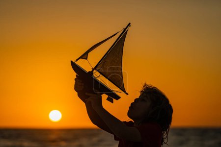 Foto de Silueta de niño jugando con el barco de vela de juguete en el mar puesta de sol. Niño disfrutando de las vacaciones de verano junto al mar. Un lindo niño jugando con un barco de juguete en la playa en un cálido día soleado de verano - Imagen libre de derechos