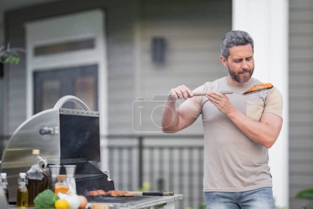 Photo for Handsome smiling hispanic man making barbecue at backyard. Picnic barbecue party. Chief cook with utensils for barbecue grill. Barbeque on holiday picnic. Man grilling a steak on BBQ - Royalty Free Image