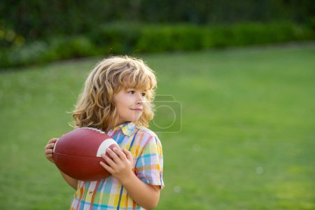 Téléchargez les photos : Enfant avec ballon de rugby. Garçon enfant avoir du plaisir et jouer au football américain sur l'herbe verte parc - en image libre de droit