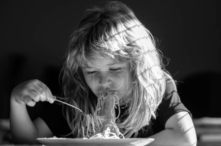 Foto de Primer plano retrato de niño divertido comiendo fideos pasta espaguetis. Niño desayunando en la cocina - Imagen libre de derechos