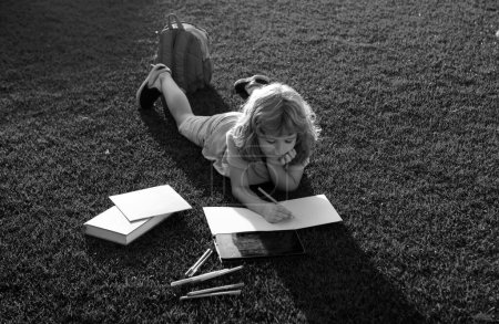 Téléchargez les photos : Joli livre de lecture garçon sur l'herbe verte notes d'écriture dans copybook. Préparation à la journée scolaire et internationale de l'alphabétisation. Concept de retour à l'école - en image libre de droit